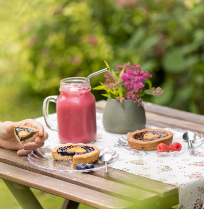 A garden table with a snack consisting of a large glass of delicious Raspberry Smoothie and two Linzer Tartelettes, Tartelette Framboise iwth raspberry and Tartelette Myrtille with blueberries.
